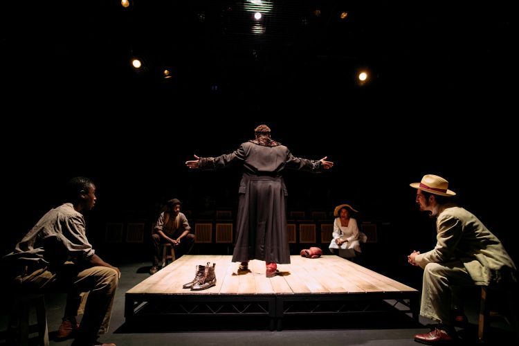 Actor with arms outstretched stands on a wooden platform, as four other actors sit at each corner of the platform